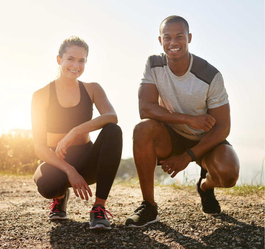 Two people in athletic wear crouch side by side on a gravel trail at sunrise, smiling toward the camera.
