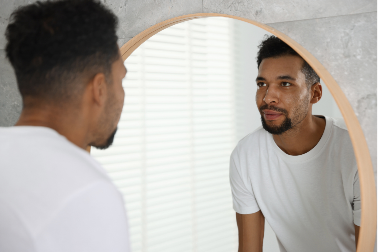 A man looks steadily at his own reflection in a round mirror.